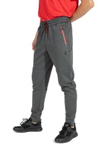 Graue Jogginghosen aus strukturiertem Stoff, mit einem verstellbaren Bund und einer Reißverschlusstasche mit roten Akzenten. Kombiniert mit schwarzen Sneakers.
