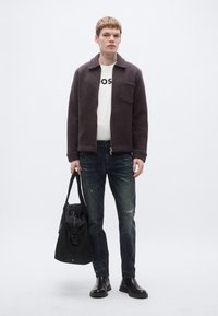 Szürke gyapjú dzseki, fehér póló "OS" felirattal, kopott kék farmer, fekete táska és fekete bokacsizma; egyszerű háttér előtt állva.