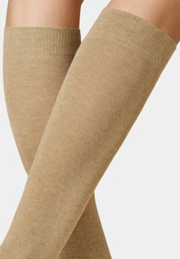 Kniestrümpfe in einem einfarbigen hellbraunen Farbton. Hergestellt aus weichem, strukturiertem Material mit einem gerippten Bündchen am oberen Ende. Glatte Oberfläche durchgängig.