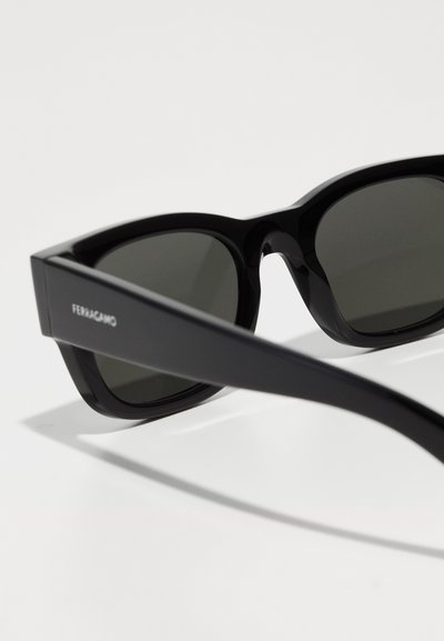 Schwarze Sonnenbrille mit einem massiven Rahmen und grünen Gläsern. Verfügt über schräg zulaufende Kanten und eine glänzende Oberfläche, die am Bügel mit "Ferragamo" markiert ist.