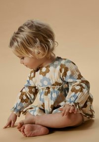 Pastellivärinen, kukkakuvioinen kevyt mekko, jossa on kerätyt hihat ja joustava vyötärö. Lapsi istuu, sormet koskettavat maata.