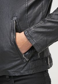 Schwarze Lederjacke mit Nahaufnahme einer Reißverschlusstasche, strukturiertem Finish, figurbetonten Ärmeln und Druckknopfverschluss an der Taille.