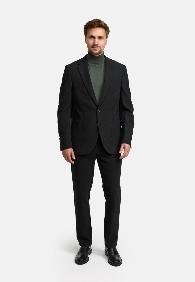 Traje negro a medida con solapa de muesca, dos bolsillos frontales y ajuste slim. Debajo, se lleva un suéter de cuello alto verde oscuro. Unos zapatos negros completan el look.