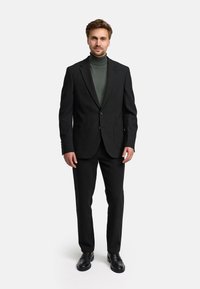 Schwarzer maßgeschneiderter Anzug mit Reverskragen, zwei Fronttaschen und schmaler Passform. Darunter wird ein dunkelgrüner Rollkragenpullover getragen. Schwarze Schuhe runden den Look ab.