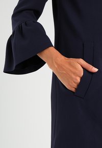 Ruka v kapse tmavě modrého oděvu s rozšířeným rukávem na světlém šedém pozadí.