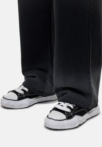 Zwarte canvas sneakers met witte rubberen zolen en neuskapjes. Voorzien van witte veters en stiksels. Draagt samen met losse, donkergrijze broeken.