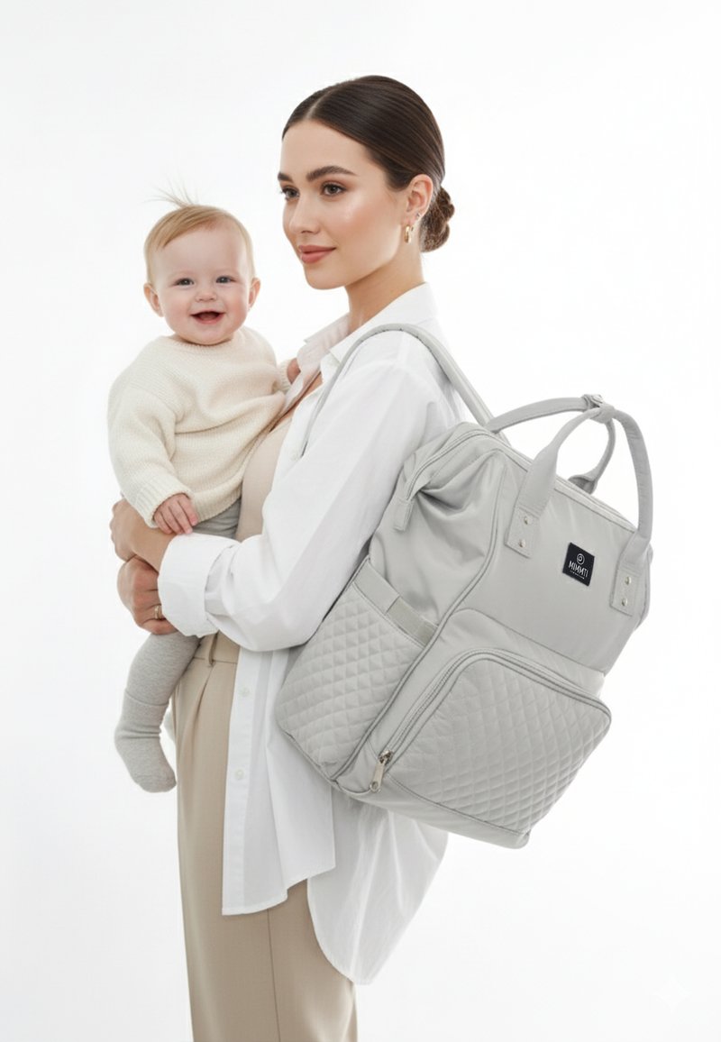 Grå quilted pusletaske med dobbelt håndtag og justerbare stropper, med en lynlåslomme og logo-tag, holdt af en person med en baby.
