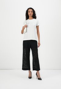 Hellgraues T-Shirt mit strukturiertem Print, kombiniert mit weiten schwarzen Spitzenhosen und schwarzen High Heels. Das Modell hält eine kleine weiße Handtasche.