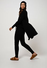 Černý pletený cardigan přes odpovídající černý outfit s kalhotami na stahování. Hnědé skládací sandály. Modelka je v pohybu na neutrálním pozadí.