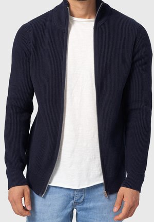 Mann trägt einen marineblauen, gerippten Reißverschluss-Pullover über einem weißen T-Shirt und hellblauen Jeans, steht vor einem einfarbigen Hintergrund.
