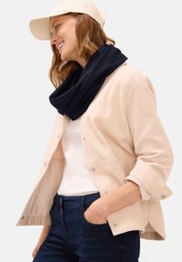 Beige kordfløjljakke med trykknapper, hvid t-shirt, mørkeblå jeans og et marineblåt tørklæde. Modellen bærer en lysefarvet kasket.