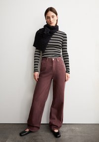 Gestreiftes schwarz-weißes Langarmoberteil, bordeauxfarbene weite Hose, schwarzer strukturierter Schal, schwarze flache Schuhe, vor einem neutralen Hintergrund.