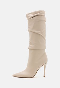 Béžová kozačka do půli stehen vyrobená z hladké kůže, s volným designem, špičatou špičkou a vysokým stiletto podpatkem se zlatým akcentem.