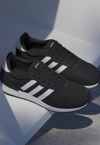 Sneakers Adidas nere con strisce bianche e suole bianche, esposte su una superficie bianca angolare che proietta ombre morbide.