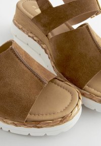 Brune ruskinds wedge sandaler med brede krydsseler, en glat tan fodseng og hvid platformssål accentueret af et vævet bånd.