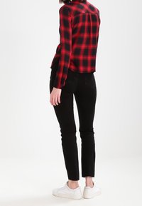 Chemise à carreaux rouge et noire avec boutons, manches longues, associée à un jean noir slim et des baskets blanches, vue de dos.