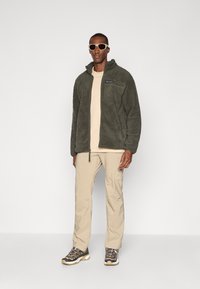 Fleecejacke in dunkelgrün mit einer Reißverschlusstasche auf der Brust und einem beigen Hemd darunter. Beiger Hosen und braune Wanderschuhe vervollständigen das Outfit.