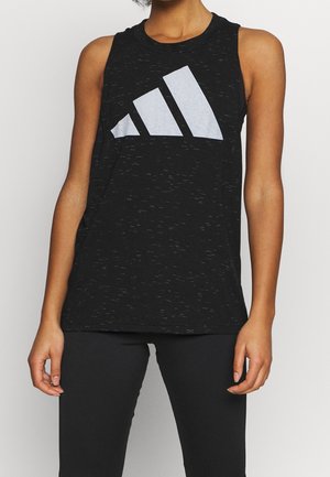 Kobieta w czarnej bluzce na ramiączkach z dużym białym logo Adidas i czarnych legginsach, stoi z rękami swobodnie opuszczonymi wzdłuż ciała.