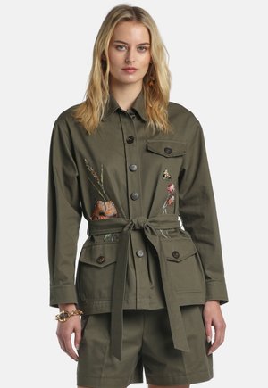 Szőke nő olíva zöld, gombos övvel ellátott dzsekit visel, virágos hímzéssel és hozzáillő rövidnadrággal, egyszínű fehér háttér előtt.