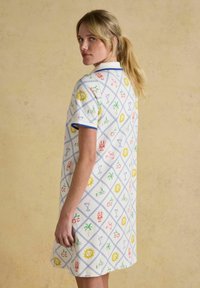 Blonde Frau mit Pferdeschwanz, die ein weißes Kleid mit blauer Borte und buntem sommerlichem Muster trägt, vor einem beigen Hintergrund steht.