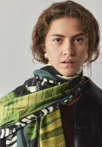 Daudzkrāsaina šalle ar zaļām, melnām un krēmkrāsas rakstiem vieglā, mīkstā audumā, brīvi apvilkta ap kaklu.