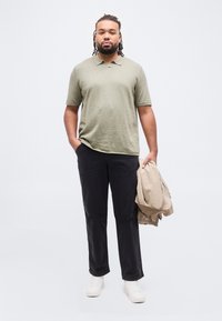 Άνδρας με πλεξούδες στα μαλλιά, φορώντας ελιάς-πράσινο πόλο, μαύρο παντελόνι και λευκά αθλητικά παπούτσια, κρατώντας ένα μπεζ σακάκι σε ένα χέρι.