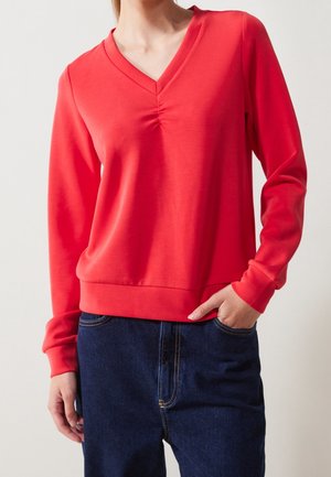 Frau trägt ein rotes Langarm-V-Ausschnitt-Sweatshirt und dunkelblaue Jeans mit einer Hand in der Tasche vor neutralem Hintergrund.