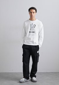 Sudadera blanca con un gráfico de una cafetera moka, con la letra negra "MOSCHINO." Pantalones cargo negros con bolsillos y zapatillas grises.