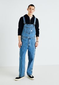 Denim overalls in light blue, featuring adjustable straps, a front pocket, and wide legs, paired with a black hooded sweatshirt and black sneakers.