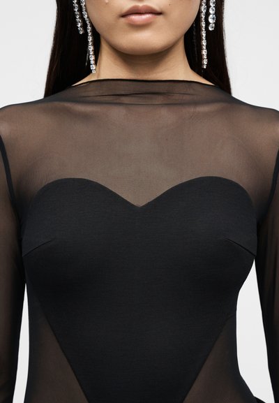 Γυναίκα φοράει μαύρο διάφανο τοπ με ντεκολτέ σε σχήμα καρδιάς και κρεμαστά σκουλαρίκια με στρας, εμφανιζόμενη από το πιγούνι έως τη μέση.