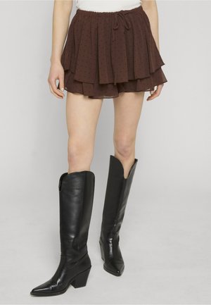 Piernas con una falda corta marrón en capas con pequeños puntos y botas negras hasta la rodilla con punta afilada y tacones gruesos.