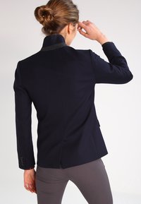 Blazer en tissu bleu marine, avec un col montant, une coupe droite et une discrète fente arrière. Le col présente un accent gris contrastant.