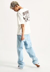 Camiseta blanca con patrón gráfico negro en la parte de atrás, combinada con vaqueros holgados de color azul claro y zapatillas negras y blancas.