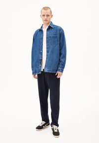 Blauwe denimshirt met een borstzak en drukknoopjes, gedragen over een lichtroze shirt; gecombineerd met donkerblauwe broek en zwarte sneakers.