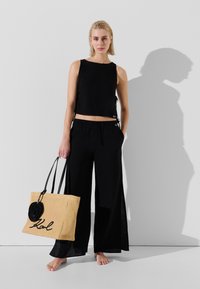 Svart kortad linne med sidoknyten, tillsammans med lösa svarta palazzo-byxor. Håller en beige vävd tote-väska med svarta logoförstärkningar.