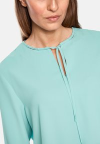 Een lichtblauwe blouse met een ronde hals, met een centrale strikaccent en zachte, gladde stof die lichtjes valt.