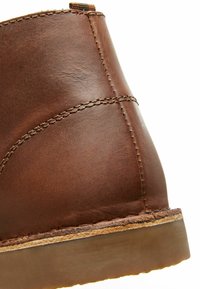 Tacón de bota de cuero marrón con una textura suave, costuras visibles y una forma redondeada. La suela de color claro añade contraste al diseño.