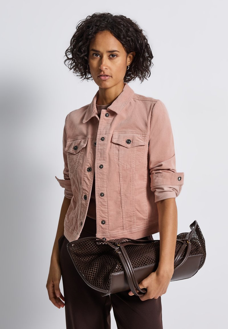 Kvinna med kort lockigt hår som bär en rosa denimjacka och håller en stor mörkbrun texturerad handväska mot en enfärgad bakgrund.