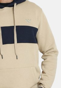Sudadera con capucha beige y azul marino con un bolsillo frontal, que presenta una franja horizontal azul marino y un logo de águila bordado en el pecho.