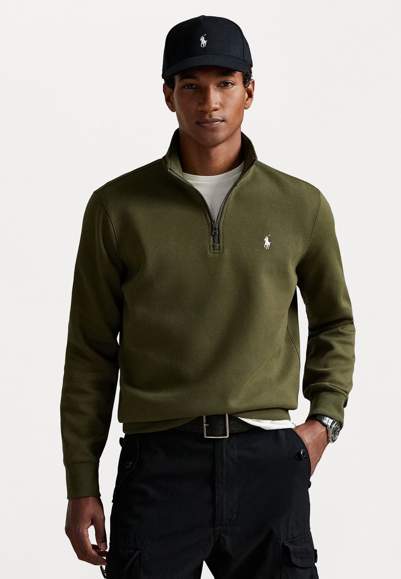 Olive green zip-neck sweatshirt with a white logo, worn over a gray t-shirt and paired with black cargo pants and a black cap.