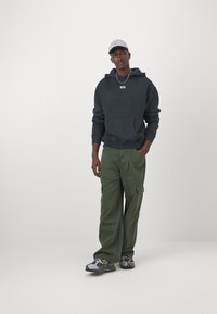 Overshirt azul escuro com bolso frontal, calças cargo verdes com múltiplos bolsos e ténis cinzentos com detalhes amarelos. Boné cinzento na cabeça.