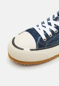 Baskets en denim avec un bout et une semelle en caoutchouc blanc, dotées de lacets blancs, d'une semelle en gomme texturée et d'accents de couture bleue.