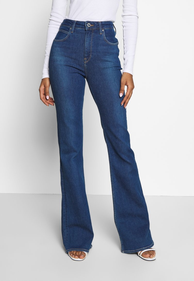 Femme portant un jean évasé bleu foncé taille haute, un haut blanc à manches longues et des sandales blanches à bout ouvert, debout devant un fond uni.