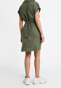 Abito camicia khaki con maniche corte e vita affusolata. Presenta un colletto a punta e un orlo arrotondato. Abbinato a sneakers bianche.