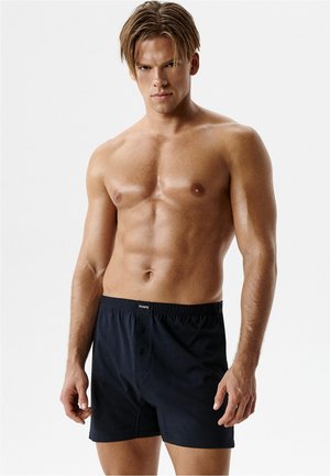 Umięśniony młody mężczyzna z krótkimi brązowymi włosami, ubrany w czarne bokserki, stoi na tle jednolitego białego tła.