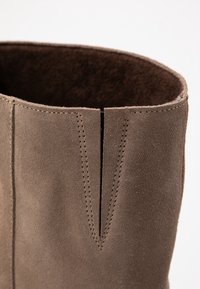 Botín de ante marrón que muestra una textura suave, cuello redondeado y una pequeña costura en forma de V en la parte frontal con puntadas en contraste.