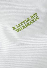 Hvid ribbet stof med grøn broderet tekst, der står "A LITTLE BIT DRAMATIC." Teksturen er subtil og struktureret.