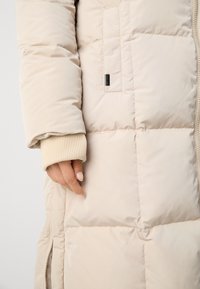 Lichtbeige pufferjack met een geribde crèmekleurige manchette, met een gewatteerde textuur, een zijzak en een merklabel nabij de zak.
