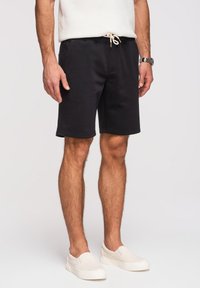Homme portant un short noir à cordon, des chaussures blanches sans lacets, un haut en maille blanc et une montre-bracelet, debout devant un fond uni.
