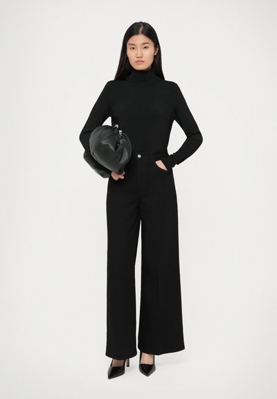 Un pulover negru cu guler înalt, asortat cu pantaloni negri largați. Modelul ține o geantă de mână rotundă din piele. Tocurile negre ascuțite completează look-ul.